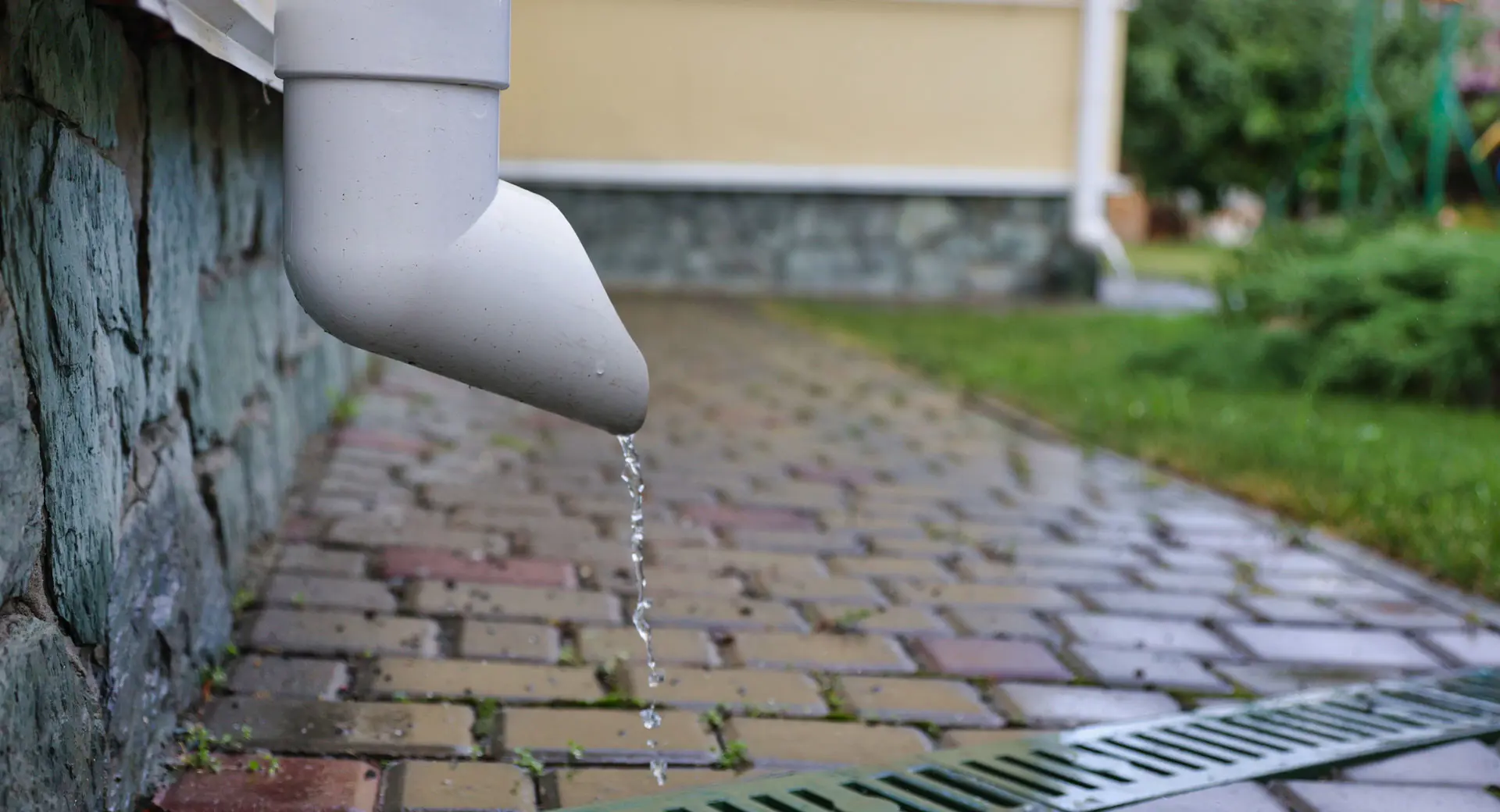 A close-up of water dripping from a house drainpipe into a drain in a paved area of a garden