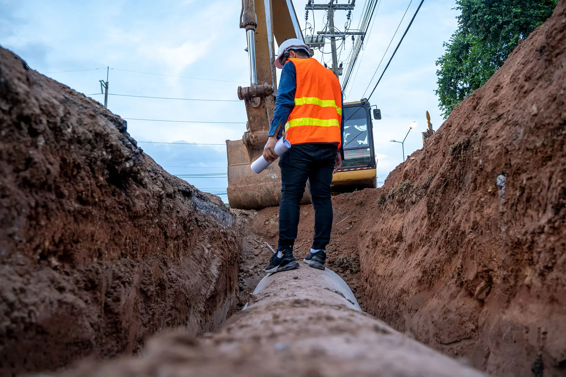 A Southern Water engineer stands on top of a pipe that has been excavated from underground. There is a digger behind them