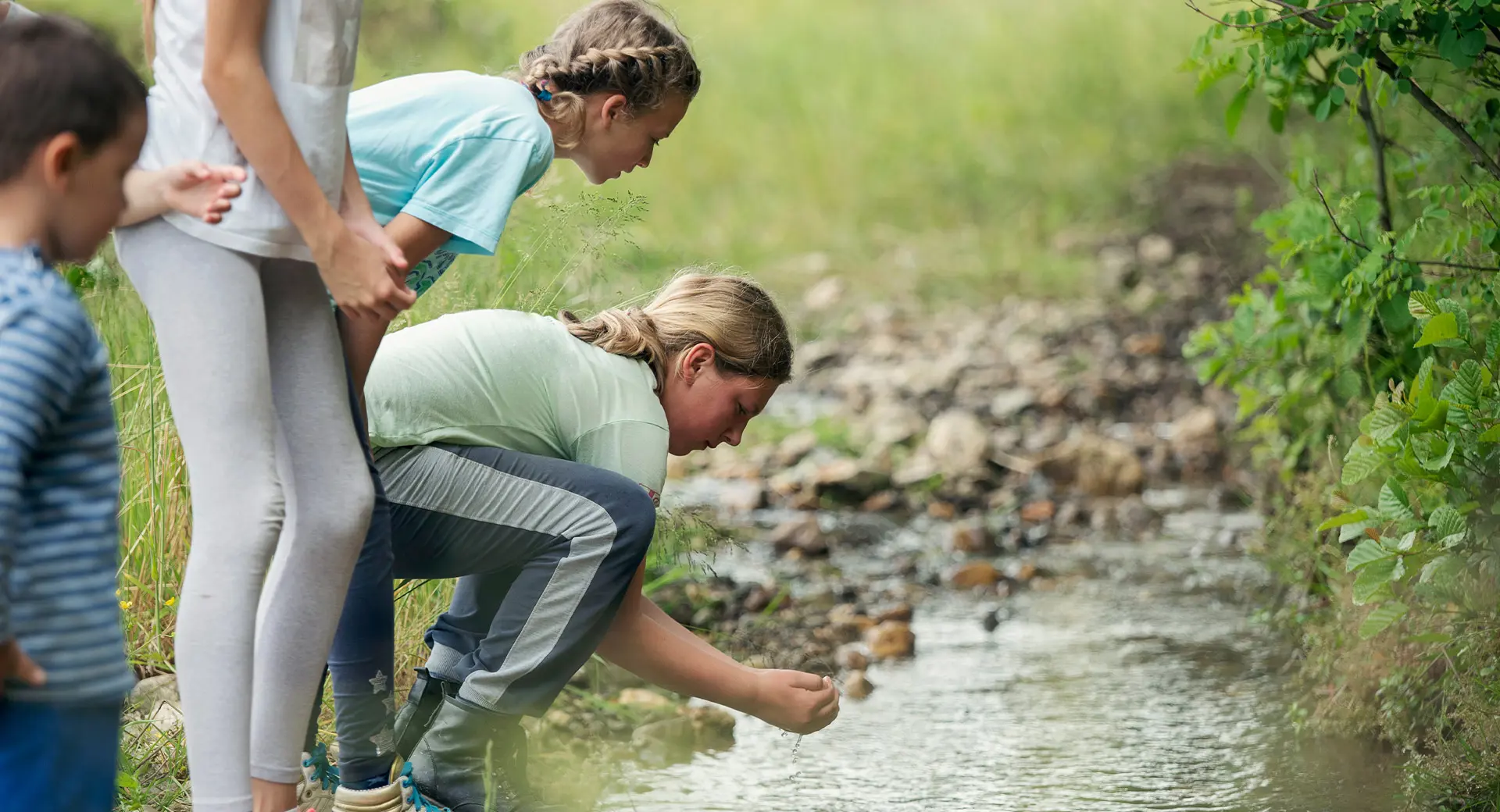 A teenager crouches and scoops water from a stream while three other children watch on